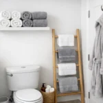 Photorealistic small modern bathroom with rolled towels on a floating shelf, a ladder rack in the corner, and a hook on the back of the door for efficient towel storage.