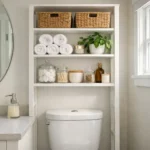 Organized small bathroom with a freestanding white storage shelf above the toilet holding baskets and towels, demonstrating space-saving solutions for apartments.